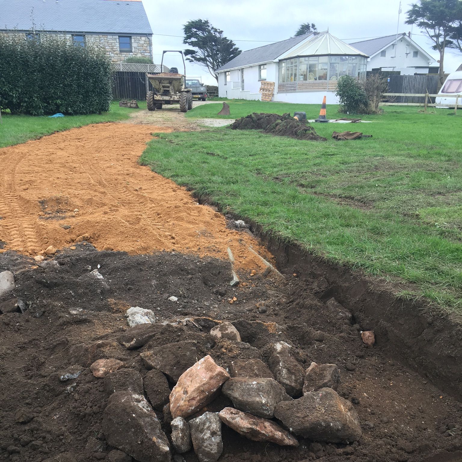 A construction site with a dirt and sand path being prepared, rocks and soil piled in the foreground, machinery and houses in the background.