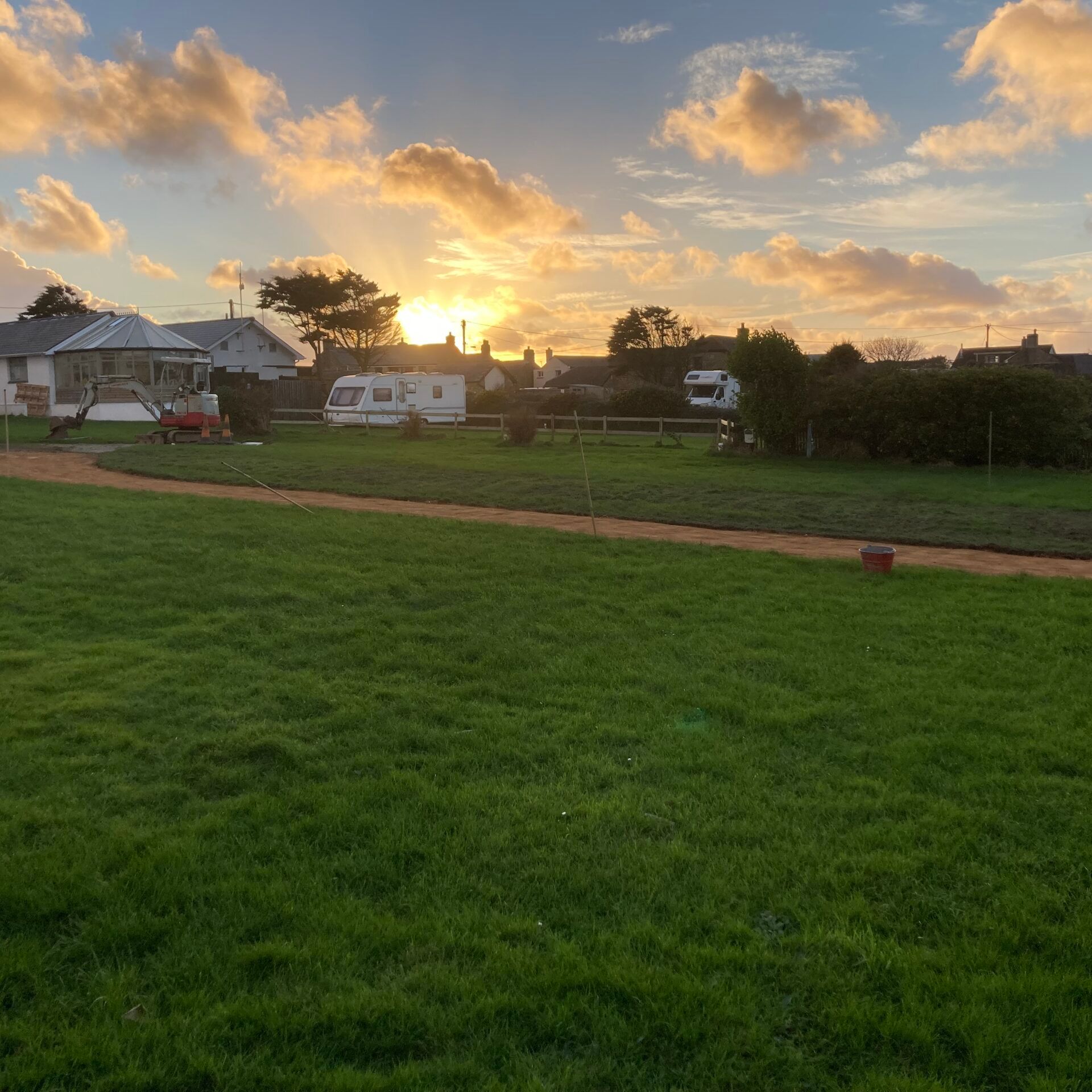 A grassy field with a dirt path leading towards houses and caravans, under a sunrise or sunset sky with scattered clouds.