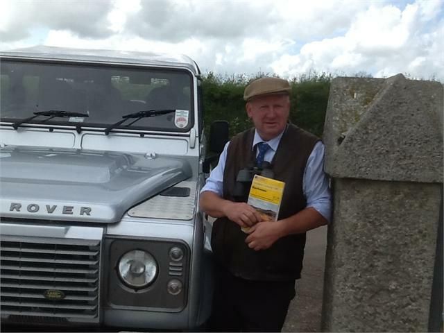 Man standing next to a silver Land Rover holding a booklet and wearing a flat cap and vest.