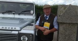 Man standing next to a silver Land Rover holding a booklet and wearing a flat cap and vest.