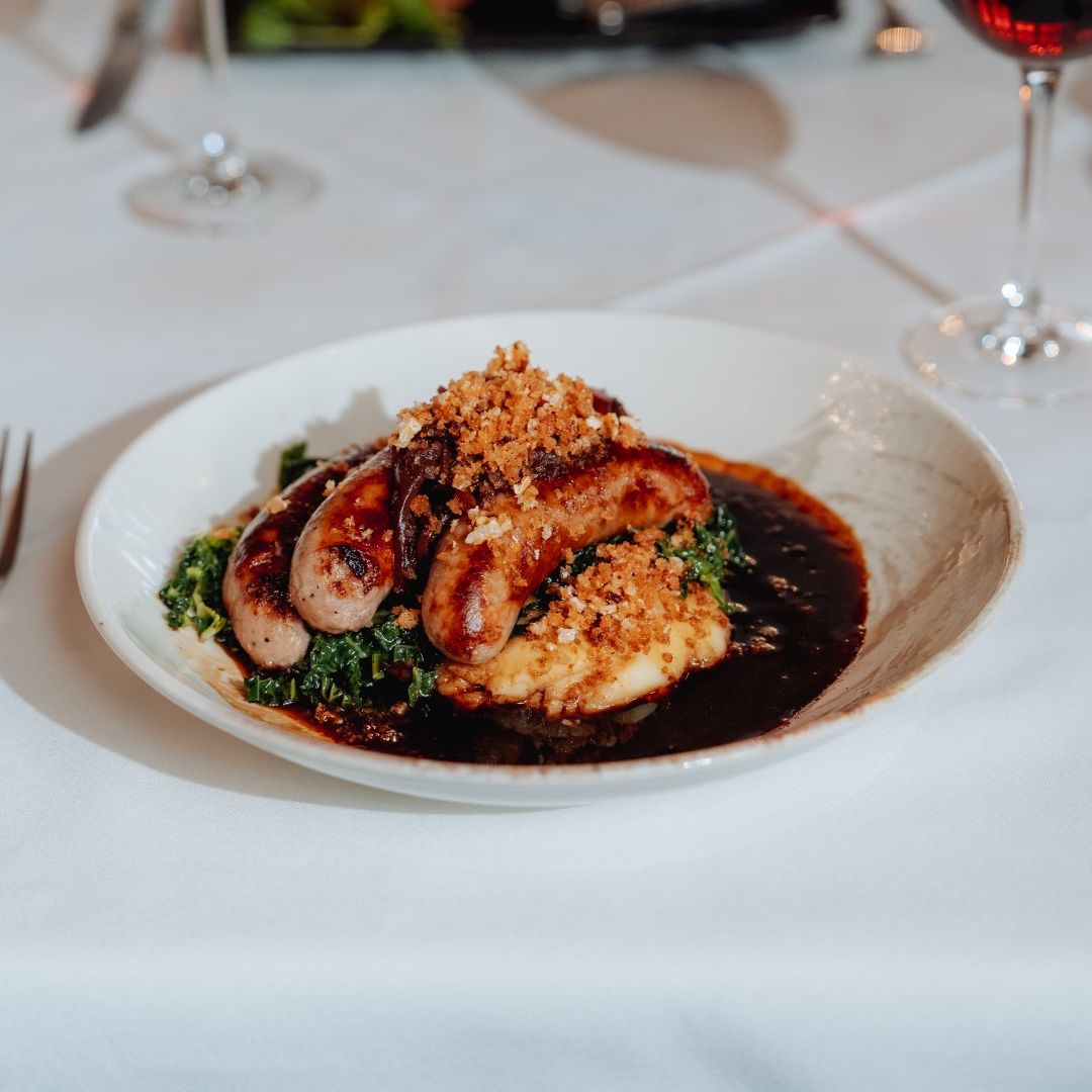 A plated dish of sausages served on mashed potatoes with greens and a rich gravy, garnished with crispy breadcrumbs.