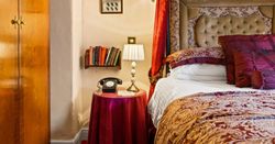 A cozy bedroom corner with a vintage telephone, table lamp, books, ornate bed, and wood wardrobe.