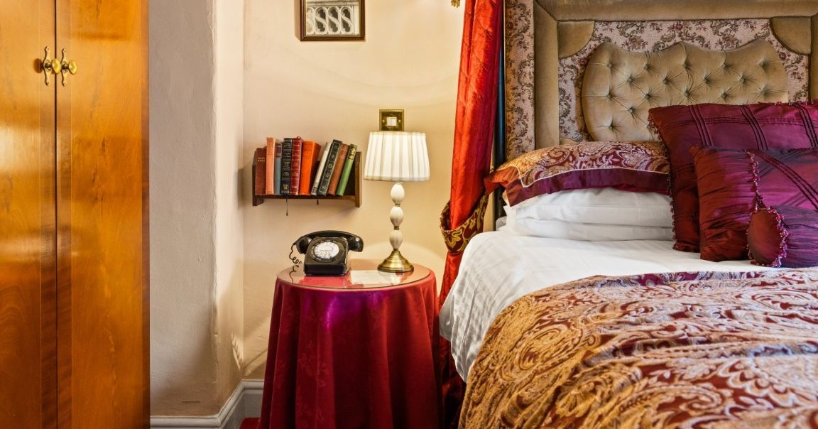 A cozy bedroom corner with a vintage telephone, table lamp, books, ornate bed, and wood wardrobe.