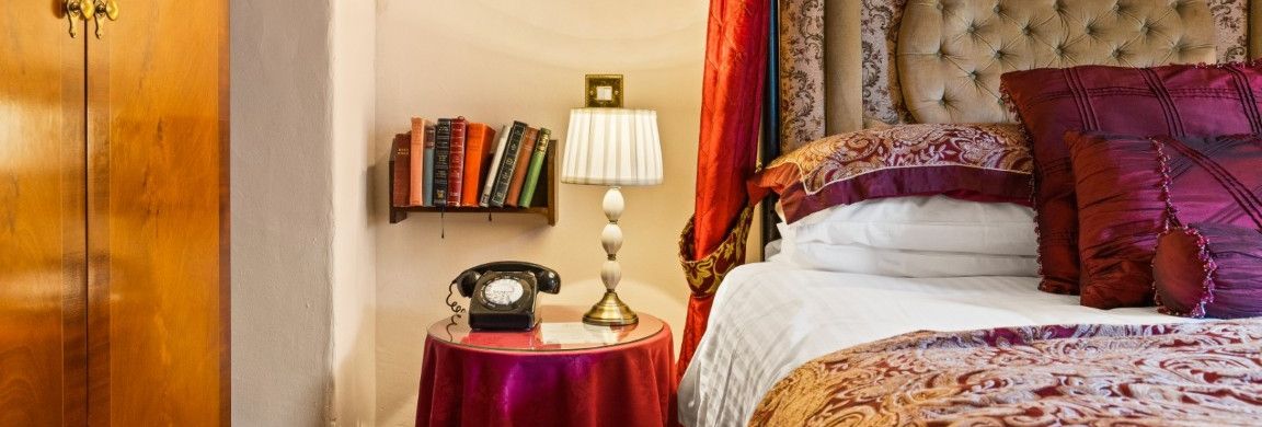 A cozy bedroom corner with a vintage telephone, table lamp, books, ornate bed, and wood wardrobe.