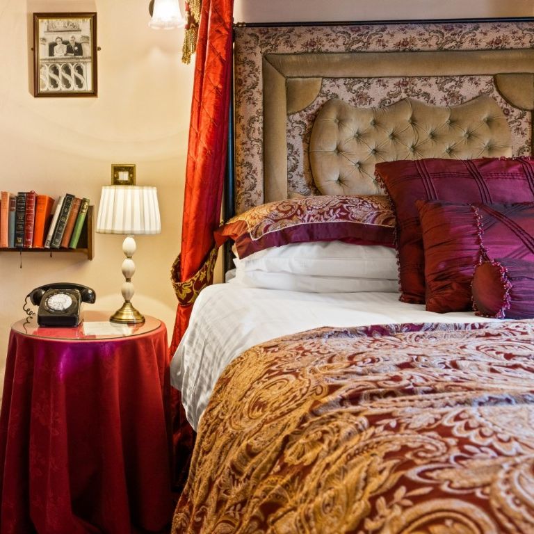 A cozy bedroom corner with a vintage telephone, table lamp, books, ornate bed, and wood wardrobe.
