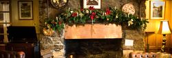 Cozy living room with a stone fireplace decorated for Christmas, featuring a copper mantel, festive garland, and vintage furniture.