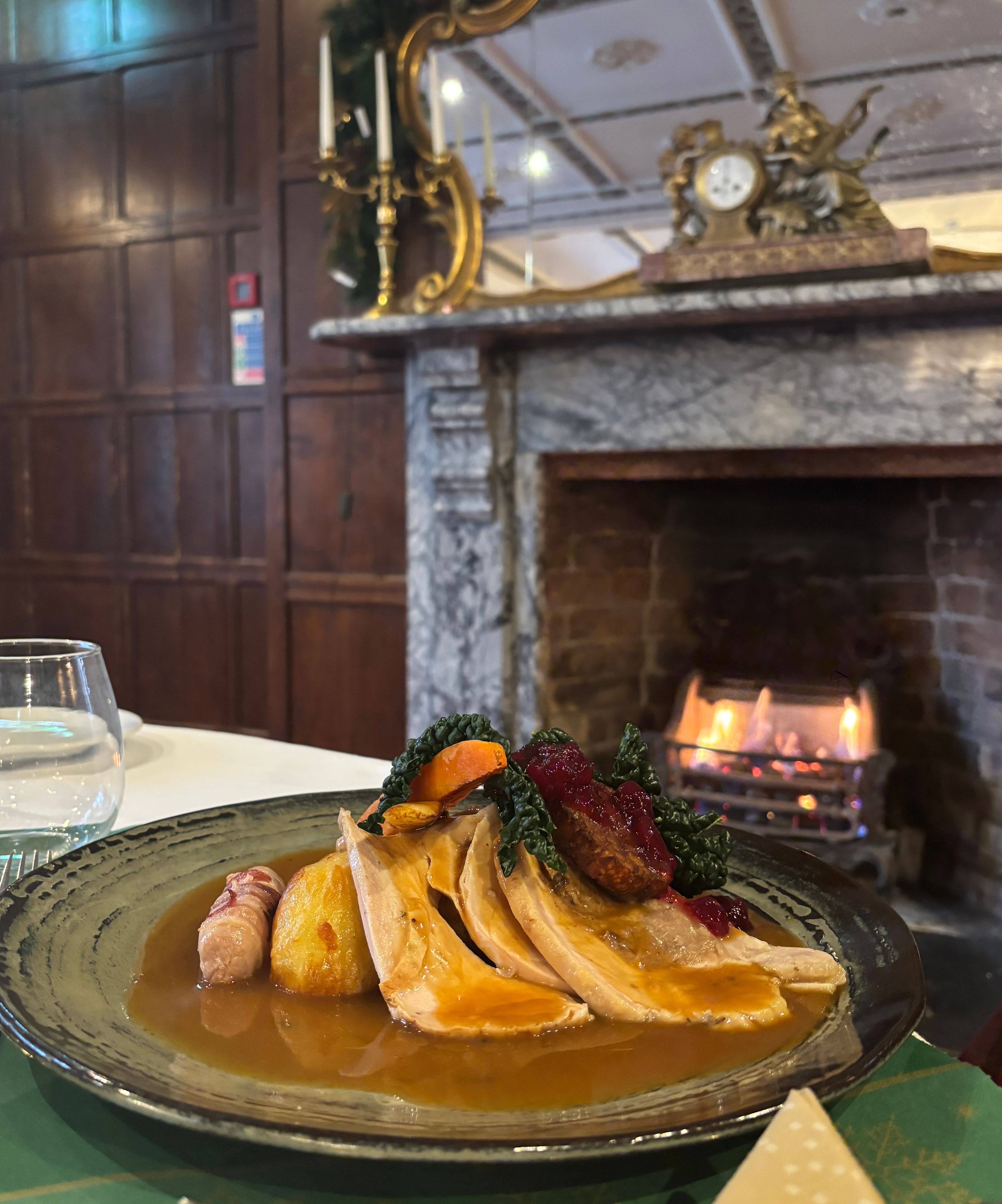 Plate of roast turkey with vegetables and sauce on a table in front of a lit fireplace in an ornate dining room.