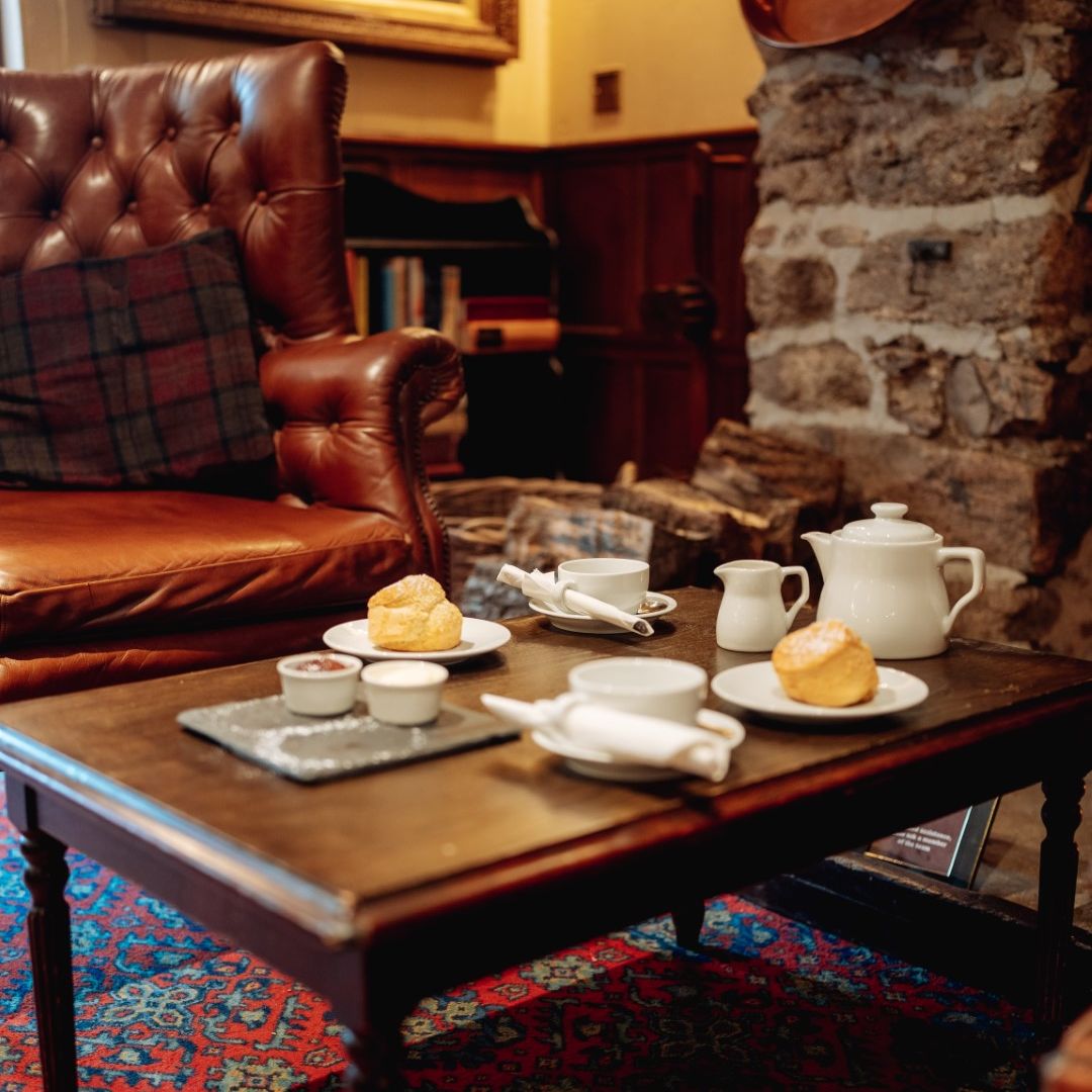 A cozy sitting area with a leather armchair and a wooden table set with scones, tea, and jam.