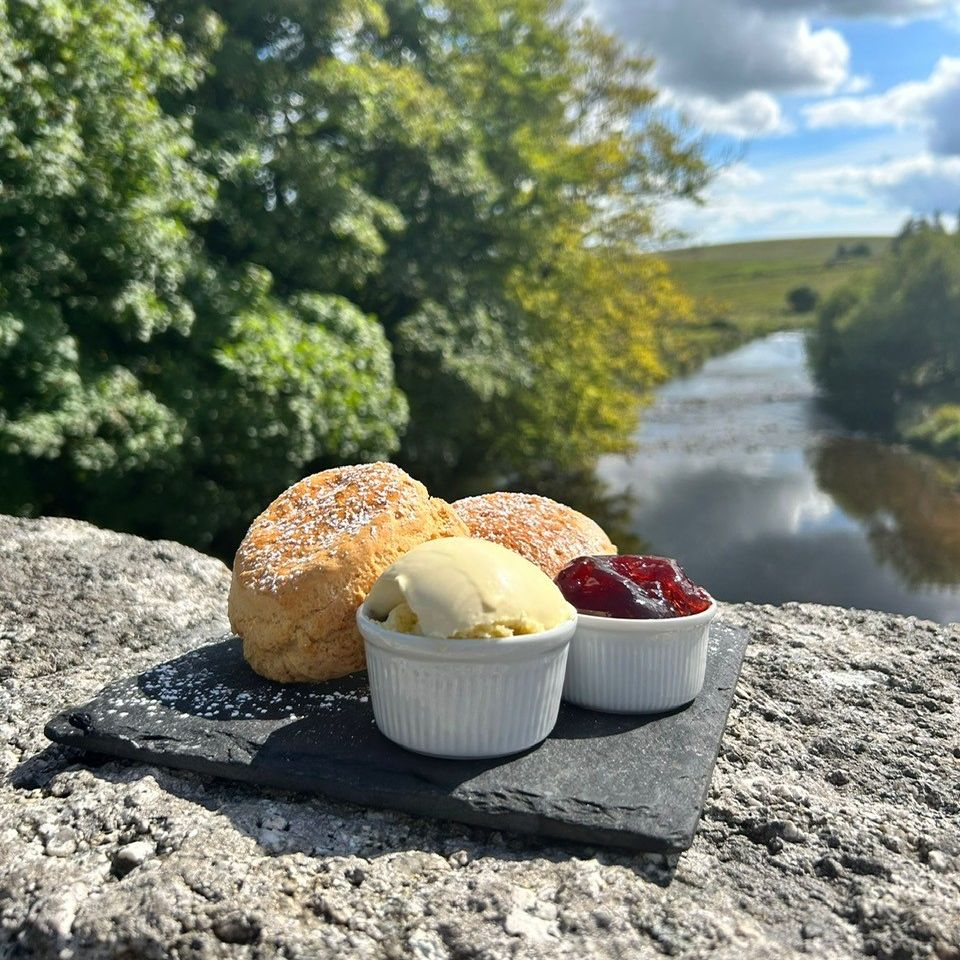 Scones with clotted cream and jam served on a slate plate on a stone bridge overlooking a scenic river and trees.