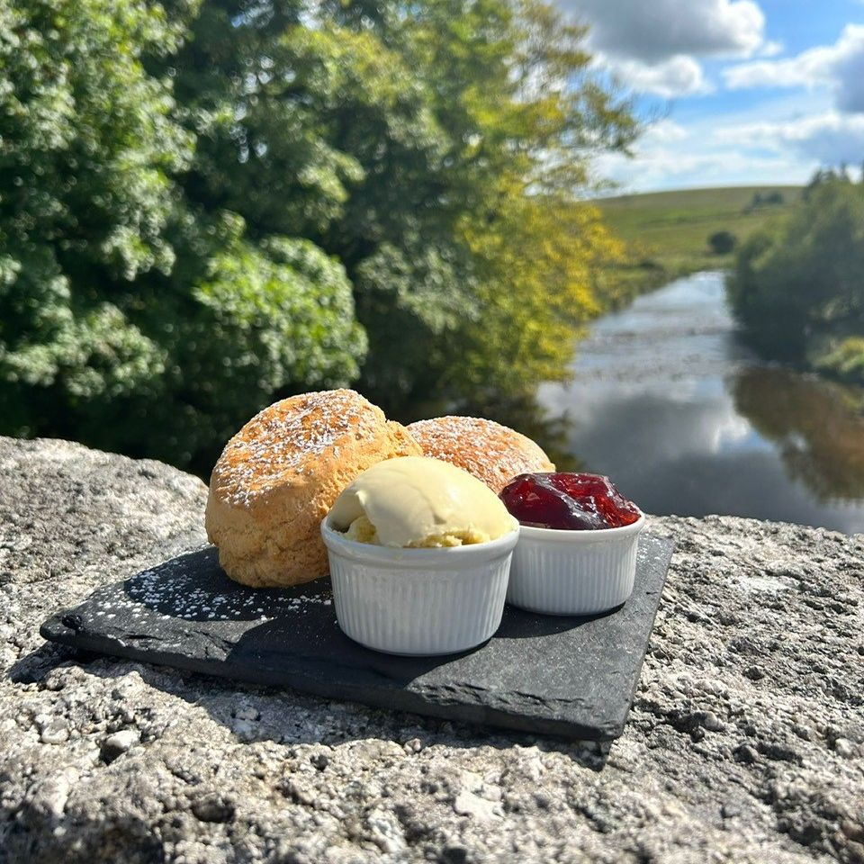 Scones with clotted cream and jam served on a slate plate on a stone bridge overlooking a scenic river and trees.