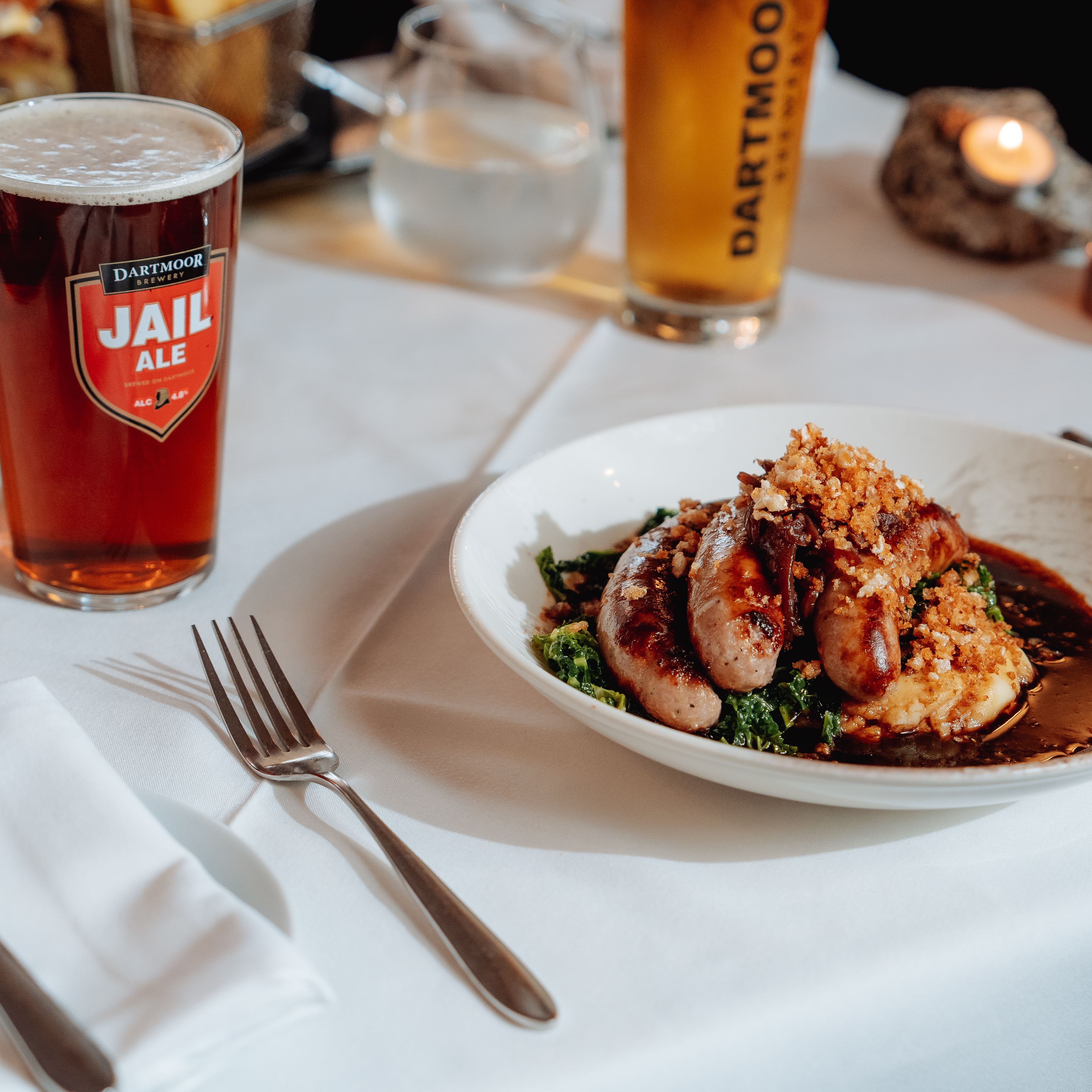 Plate of sausages and mashed potatoes served with a pint of Dartmoor Jail Ale on a restaurant table.