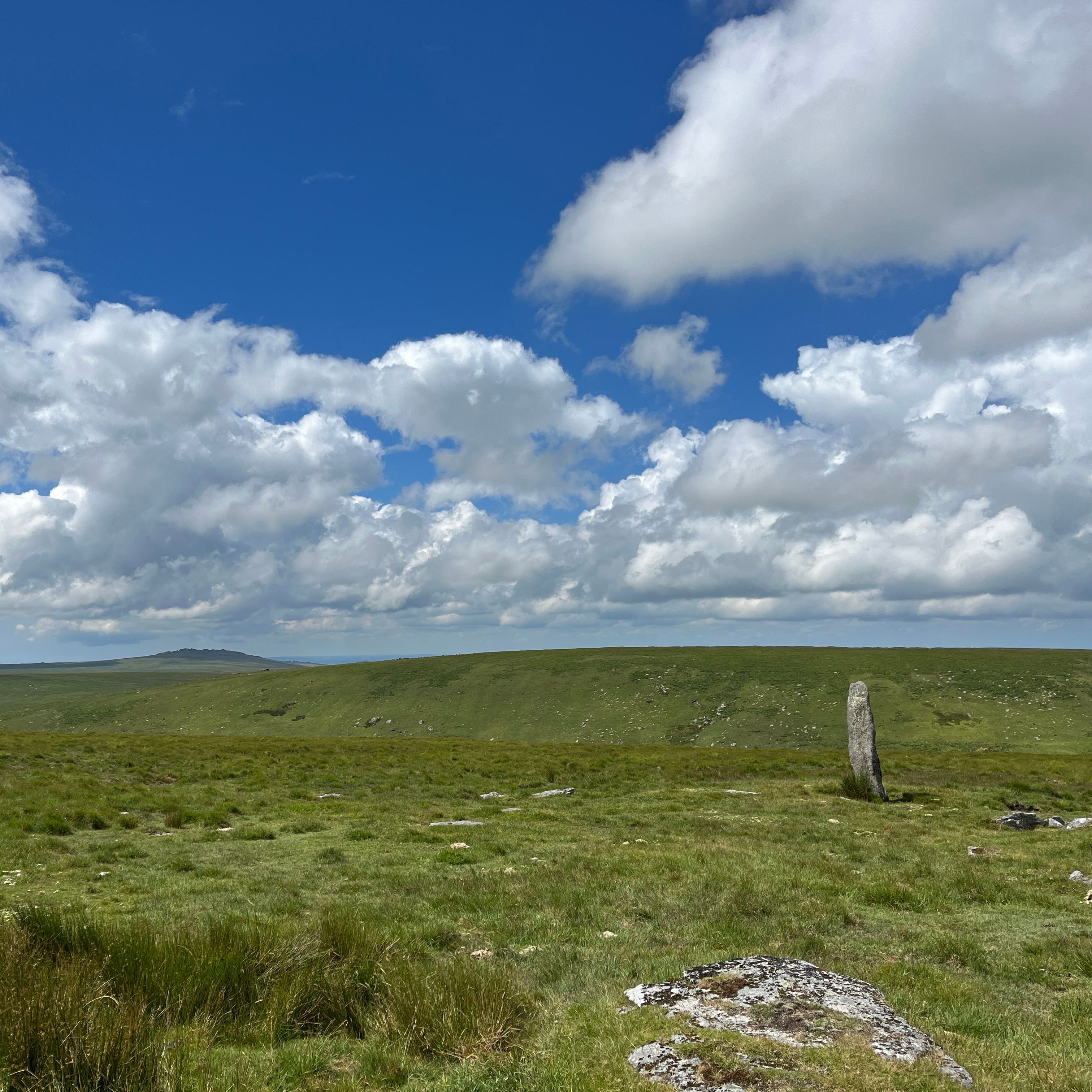 Single standing stone on a grassy moorland under a bright blue sky with scattered clouds.