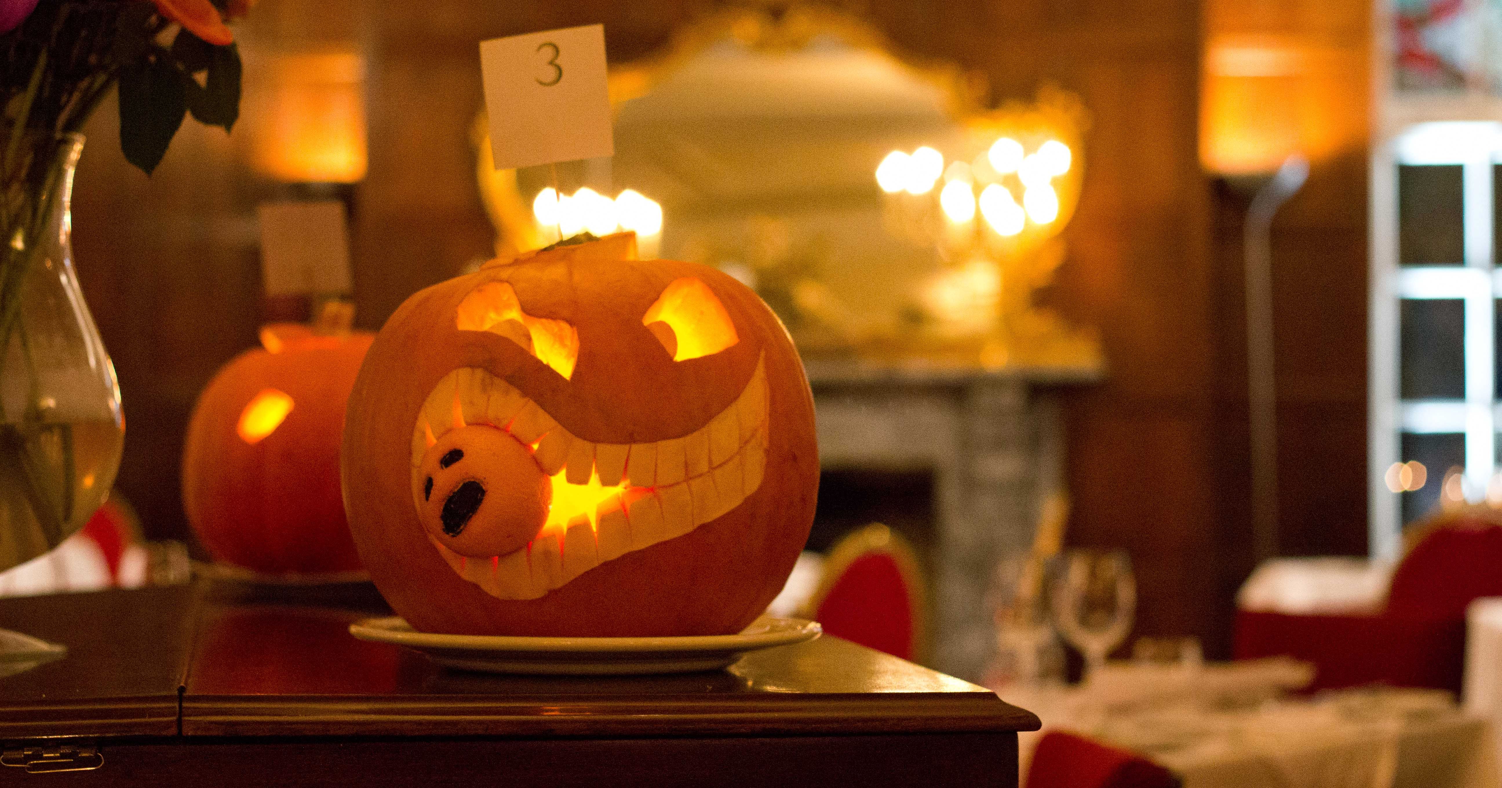 Carved pumpkin with a toothy grin and a smaller pumpkin inside its mouth, displayed indoors with a table number card and a blurred background.