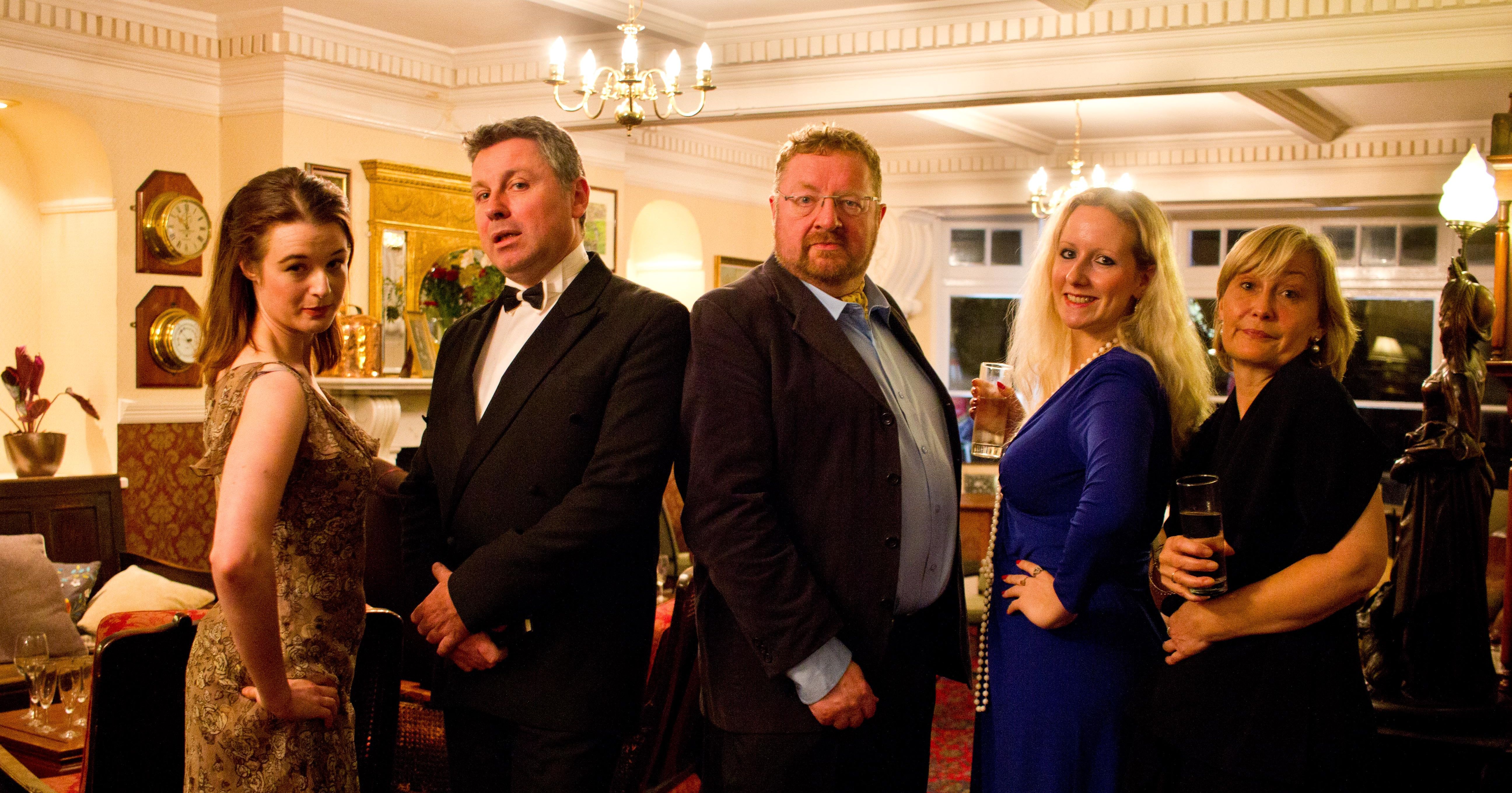 Five adults dressed formally standing in a line in an elegant, vintage-style room.