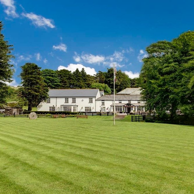 Large white country house with green lawn and surrounding trees under a blue sky.