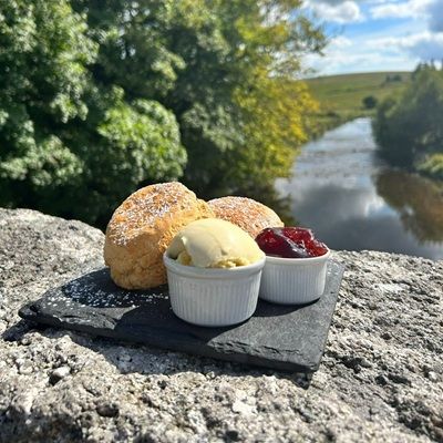 Scones with cream and jam served on a slate plate atop a stone bridge with a scenic river in the background.