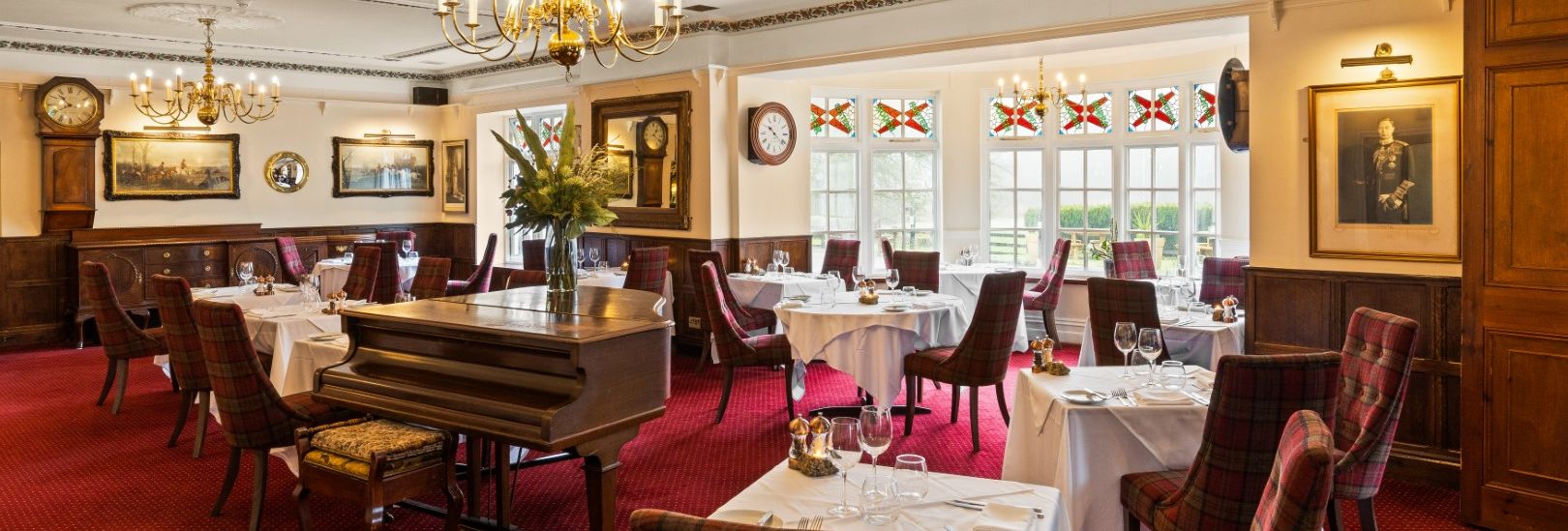 Elegant restaurant dining room with red carpet, chandeliers, tartan chairs, and a grand piano.