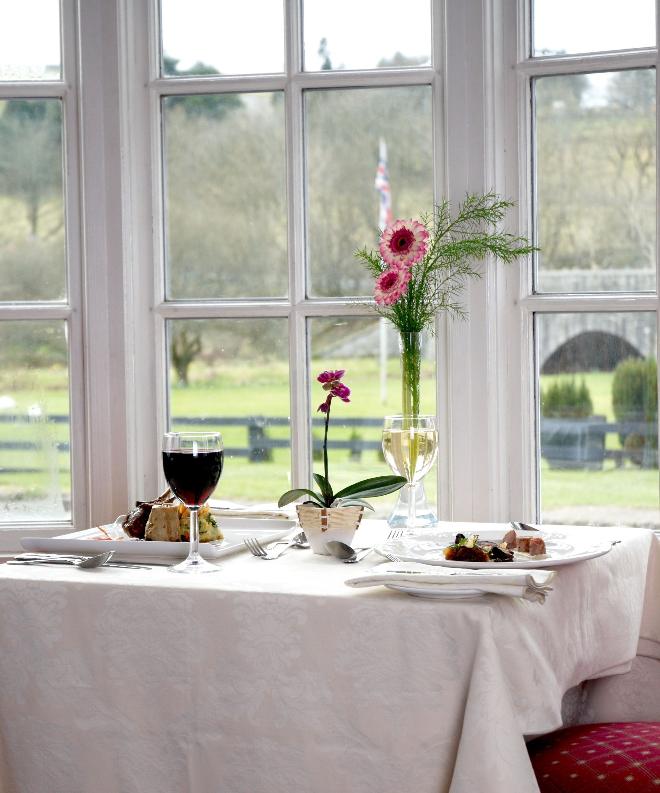 A table set for two by a large window, with red chairs, plates of food, glasses of wine, and flowers in vases.