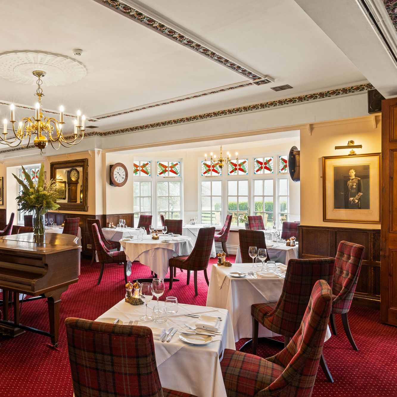 Elegant restaurant dining room with red plaid chairs, white tablecloths, chandeliers, and a grand piano.