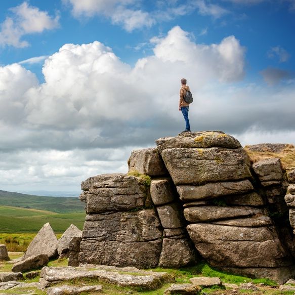 Person standing on a large rocky tor overlooking a green landscape under a partly cloudy sky.