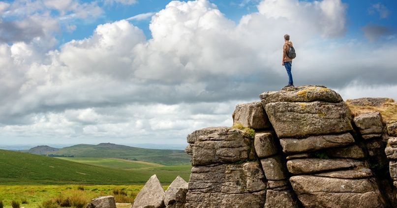 Person standing on a large rocky tor overlooking a green landscape under a partly cloudy sky.