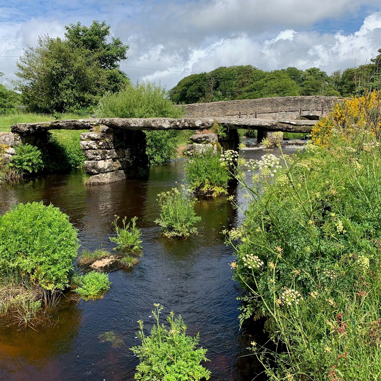 Ancient stone clapper bridge and modern bridge at Postbridge over a shallow river surrounded by lush greenery.