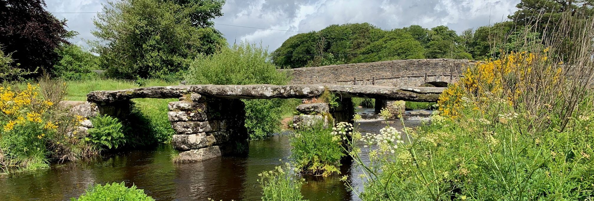 Ancient stone clapper bridge and modern bridge at Postbridge over a shallow river surrounded by lush greenery.