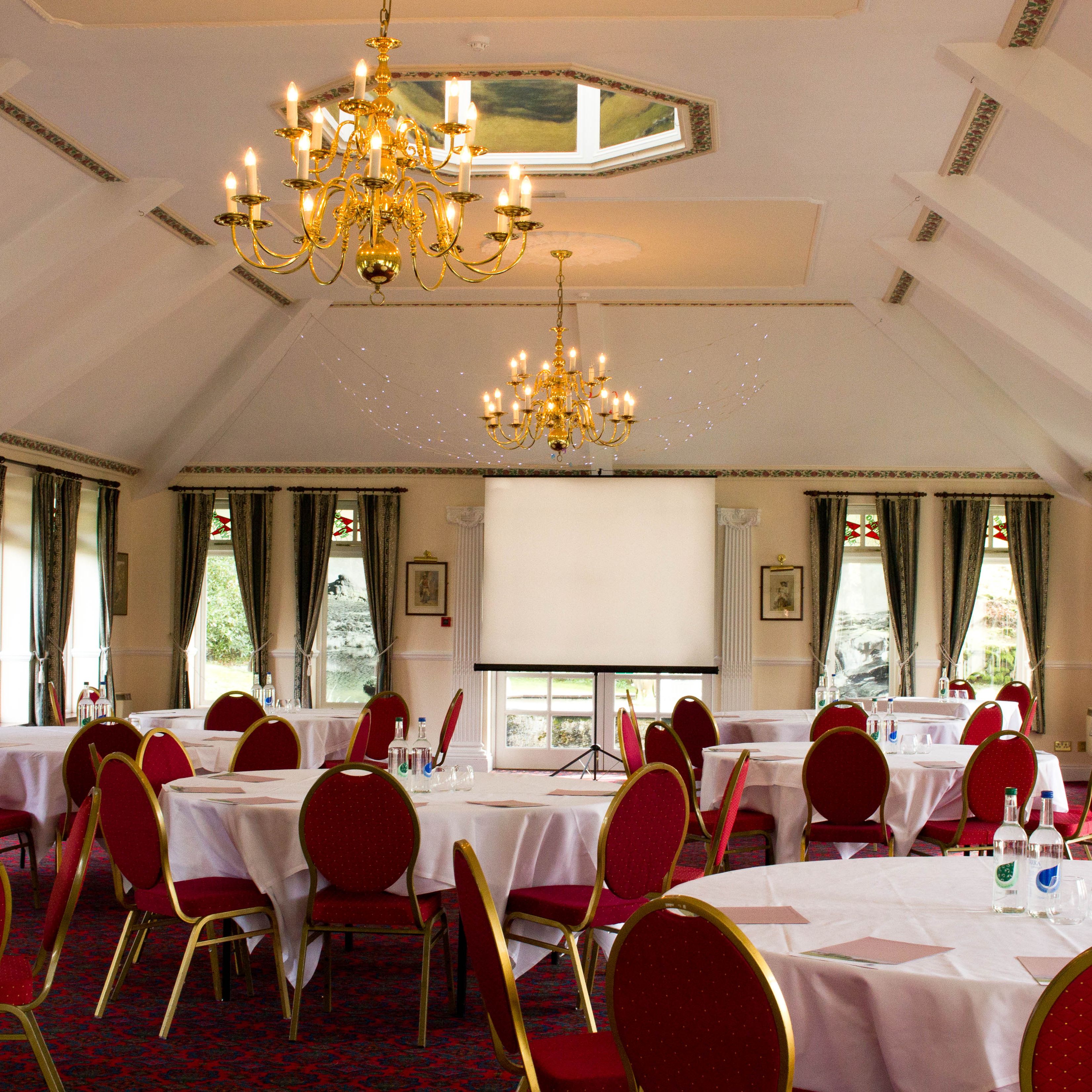 Elegant conference room with round tables covered in white cloths, red chairs, chandeliers, and a projection screen.