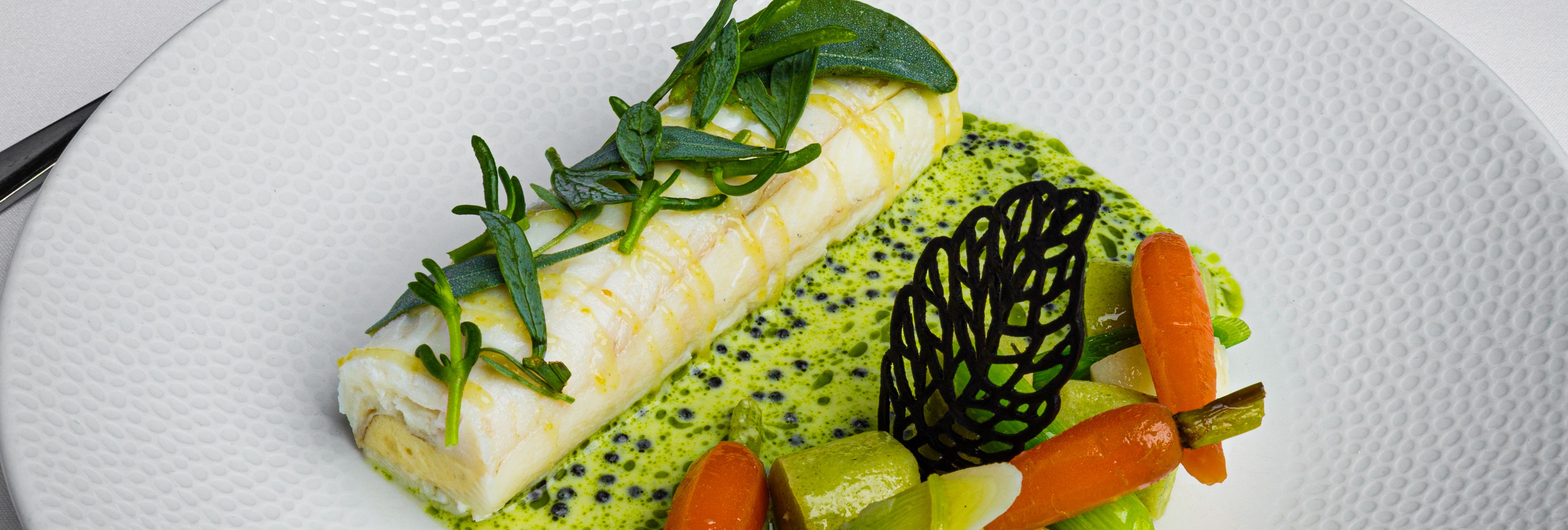 Gourmet seafood dish with vegetables and green sauce on a white plate, set on a restaurant table.