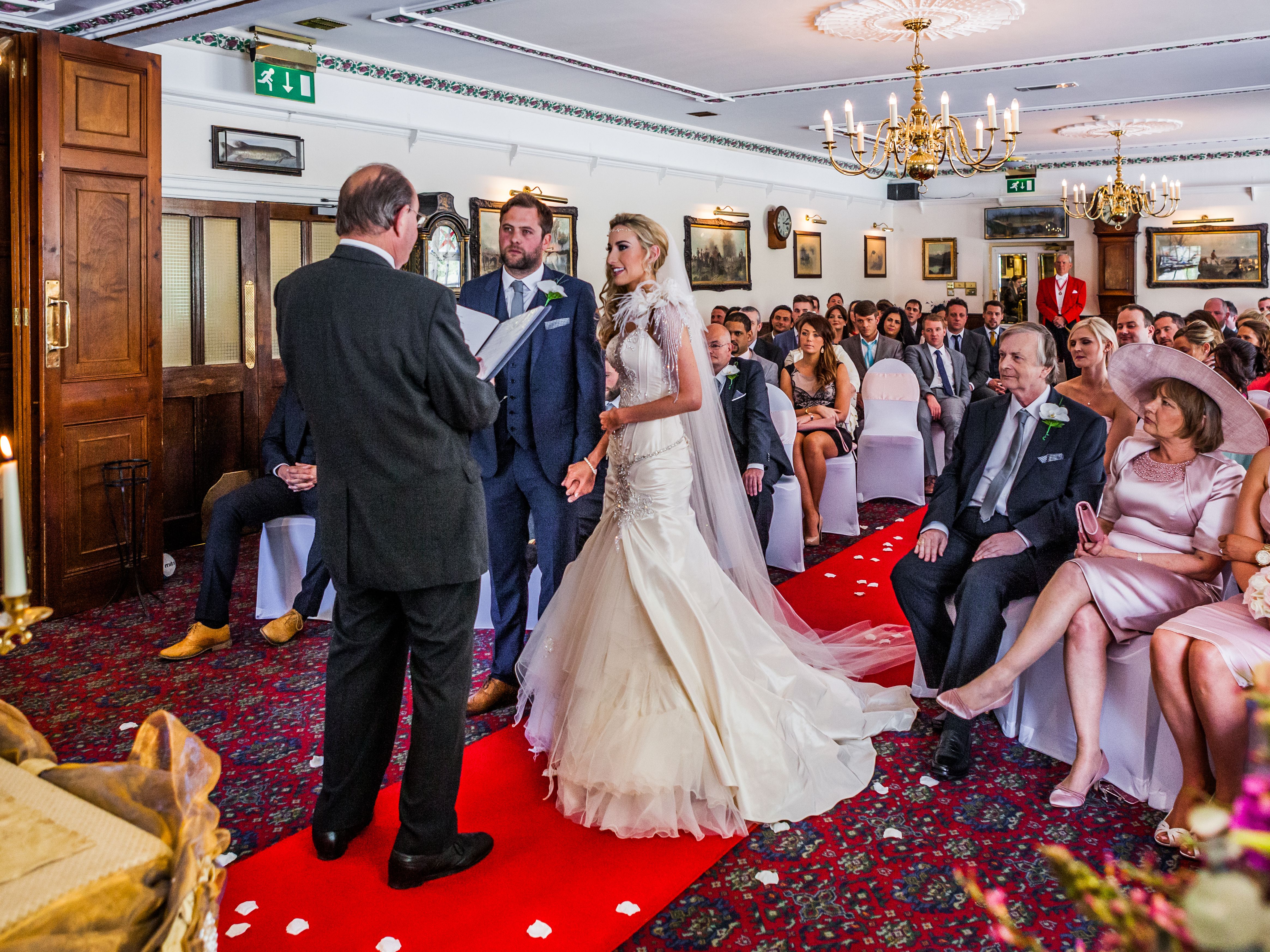 Bride and groom standing at the altar during a wedding ceremony, surrounded by guests in a formal indoor setting.