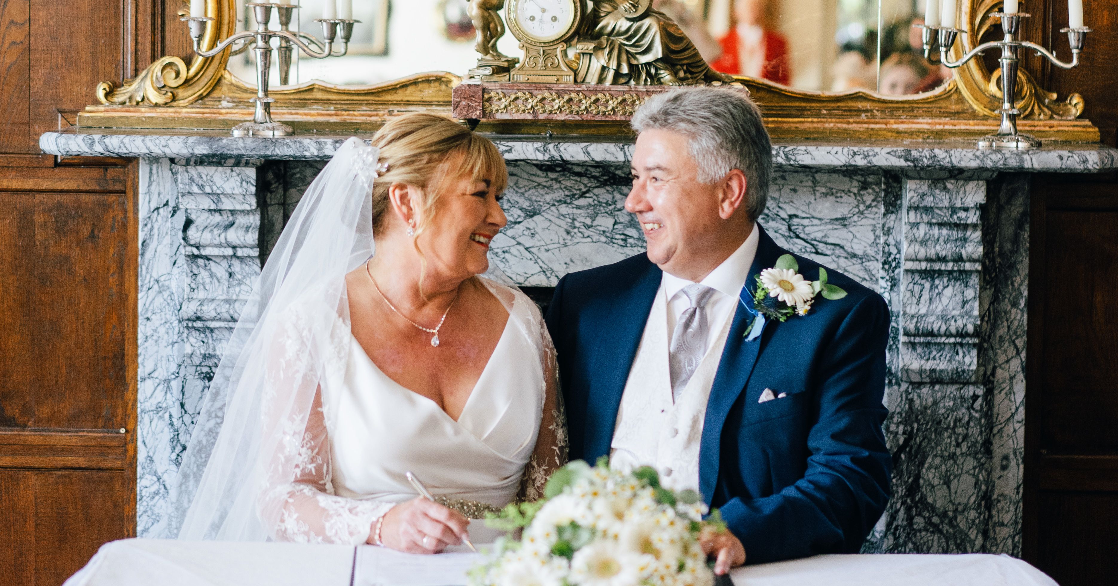Bride and groom smiling at each other while signing a document at their wedding ceremony table.