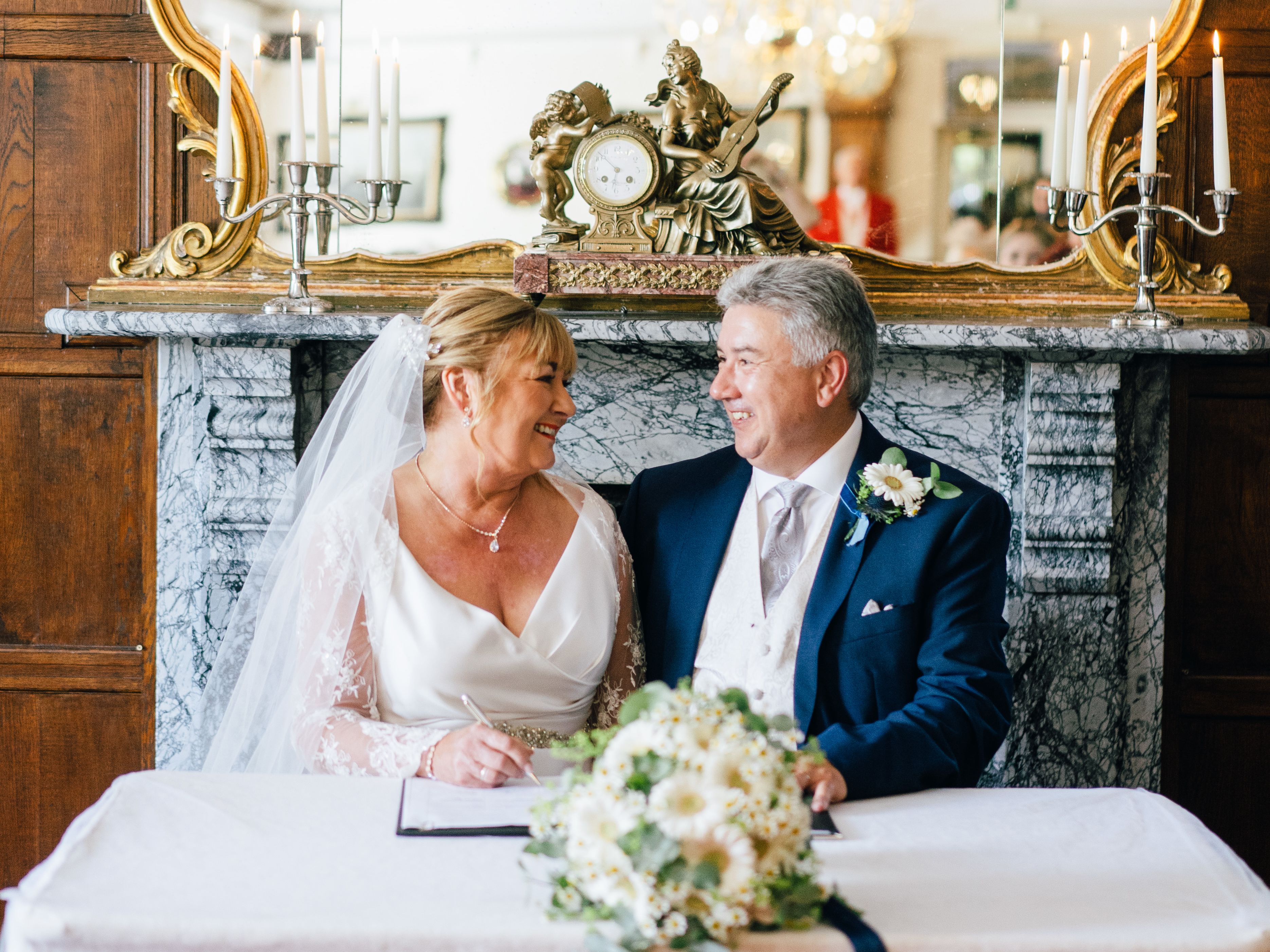 Bride and groom smiling at each other while signing a document at their wedding ceremony table.