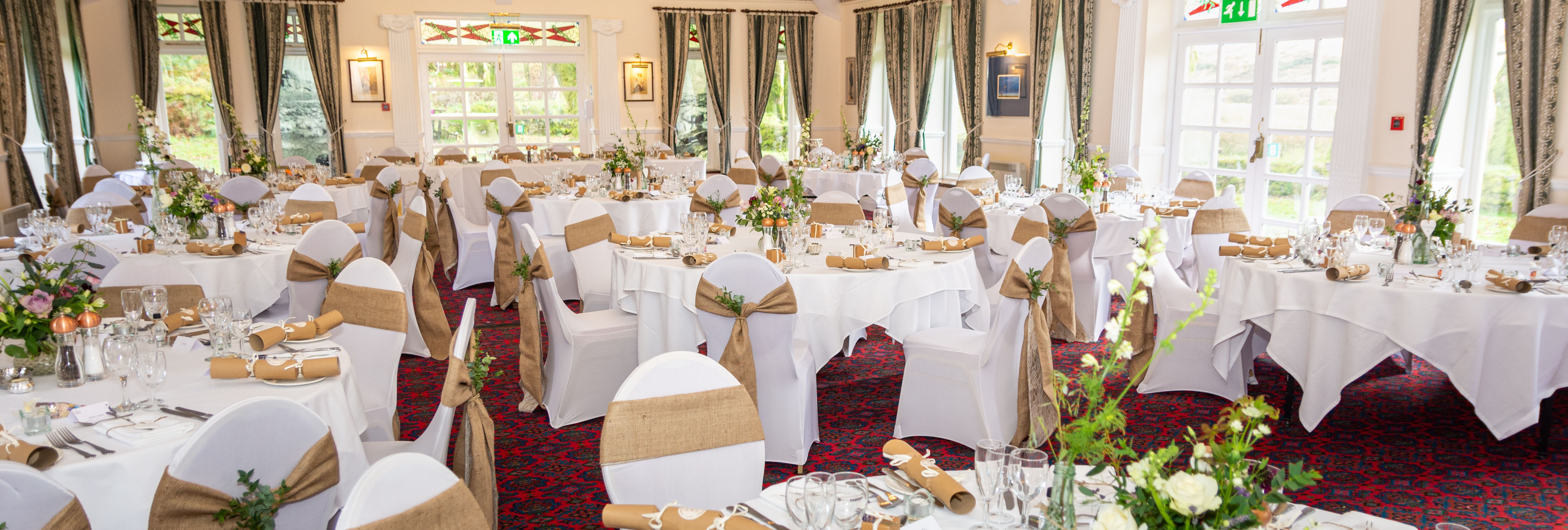 Elegant wedding reception room with round tables dressed in white cloth, rustic chair sashes, and floral centerpieces.