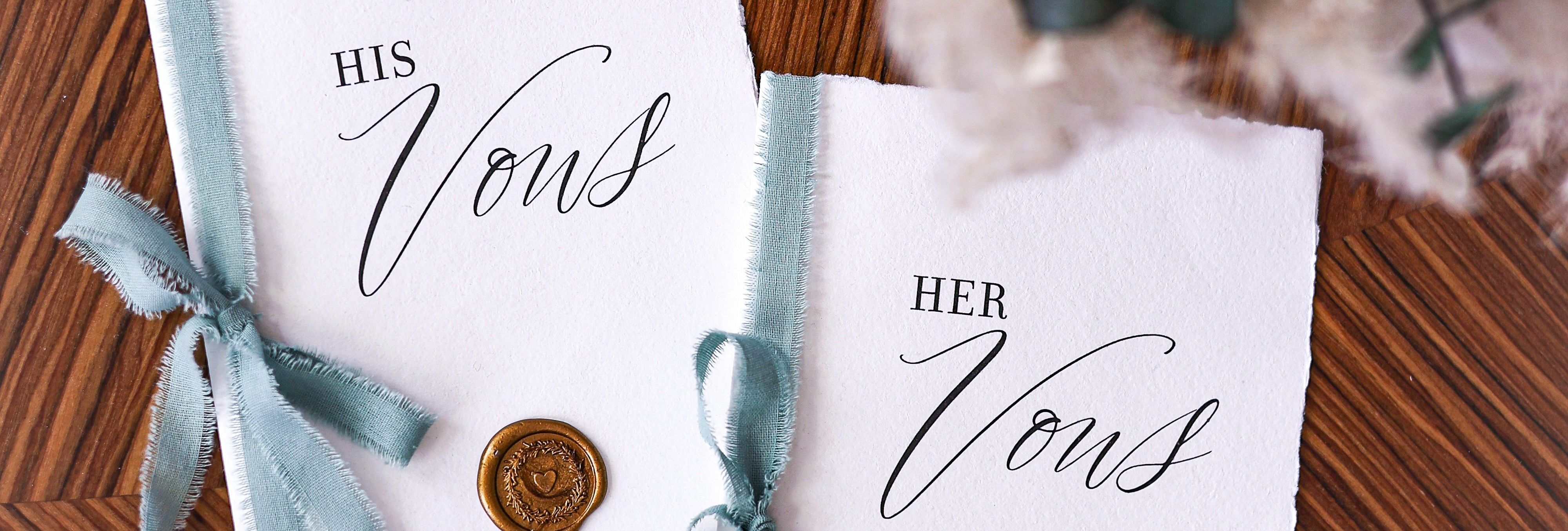 His and her wedding vow booklets with blue ribbons and wax seals next to a bouquet of flowers on a wooden table.