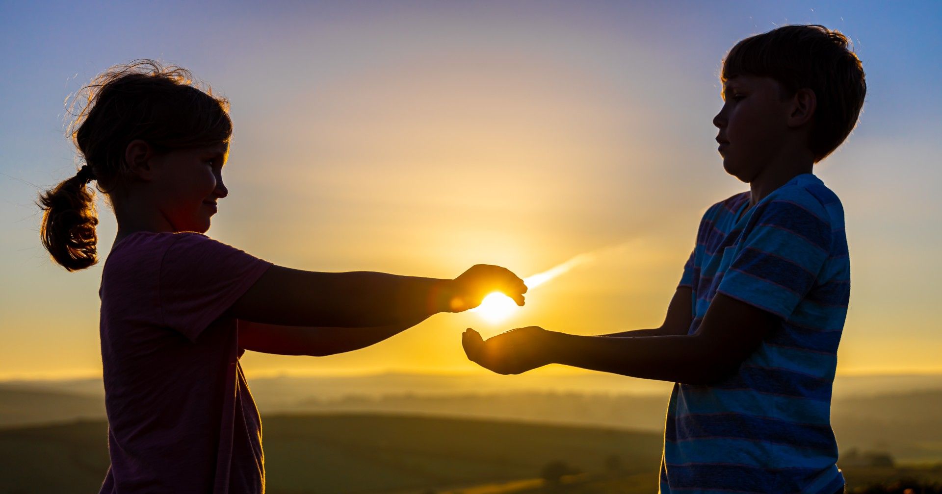 Two children silhouetted against the sunset, appearing to hold the sun between their hands.
