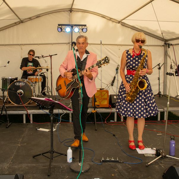A four-piece band performing on stage under a white tent. The musicians are playing electric guitar, bass, drums, and a saxophone.