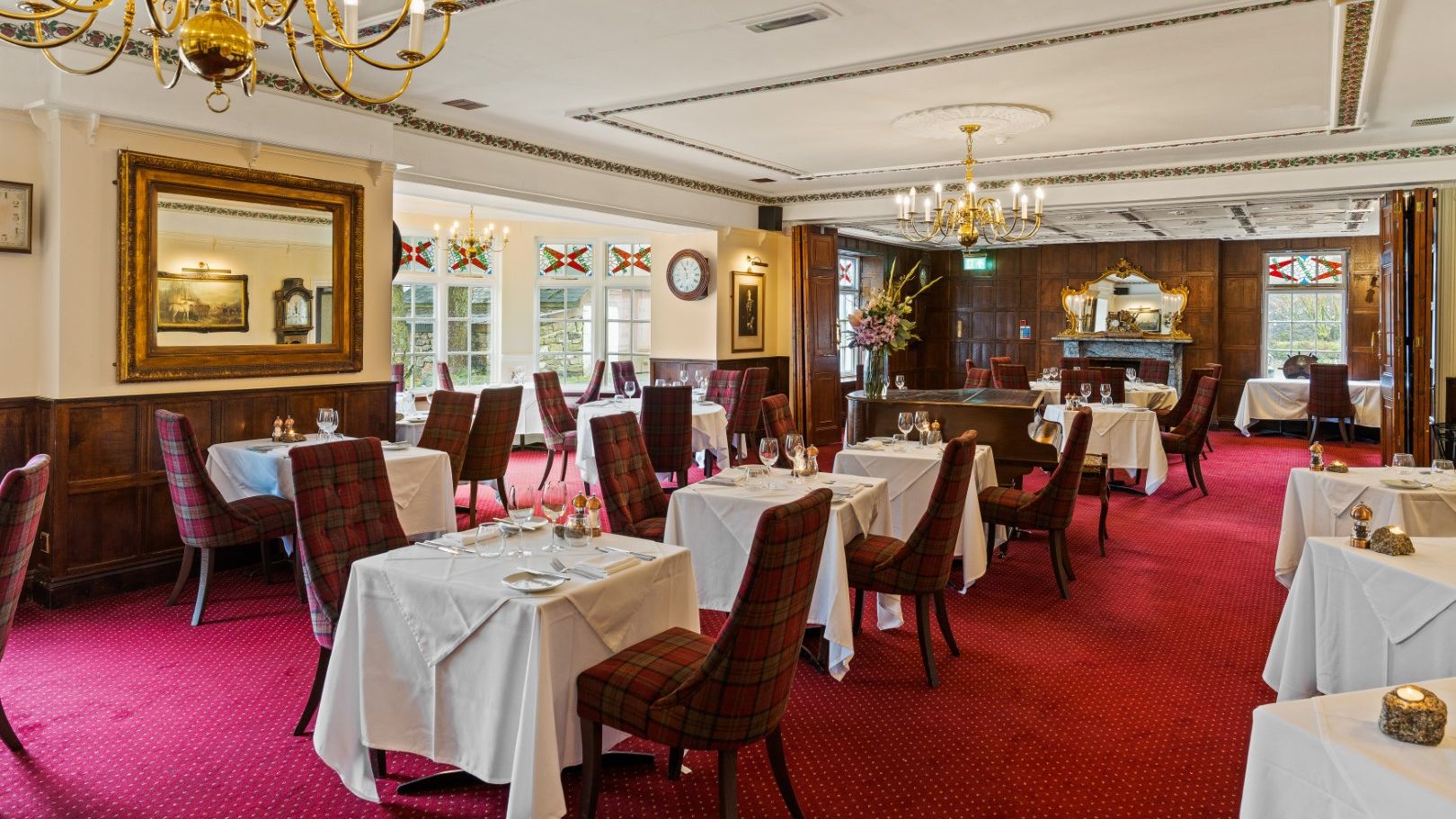 Elegant restaurant dining room with red carpet, white tablecloths, plaid chairs, chandeliers, and wooden paneling.