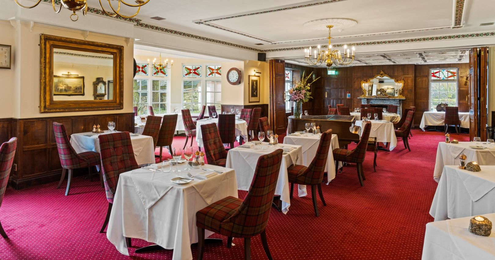 Elegant restaurant dining room with red carpet, white tablecloths, plaid chairs, chandeliers, and wooden paneling.