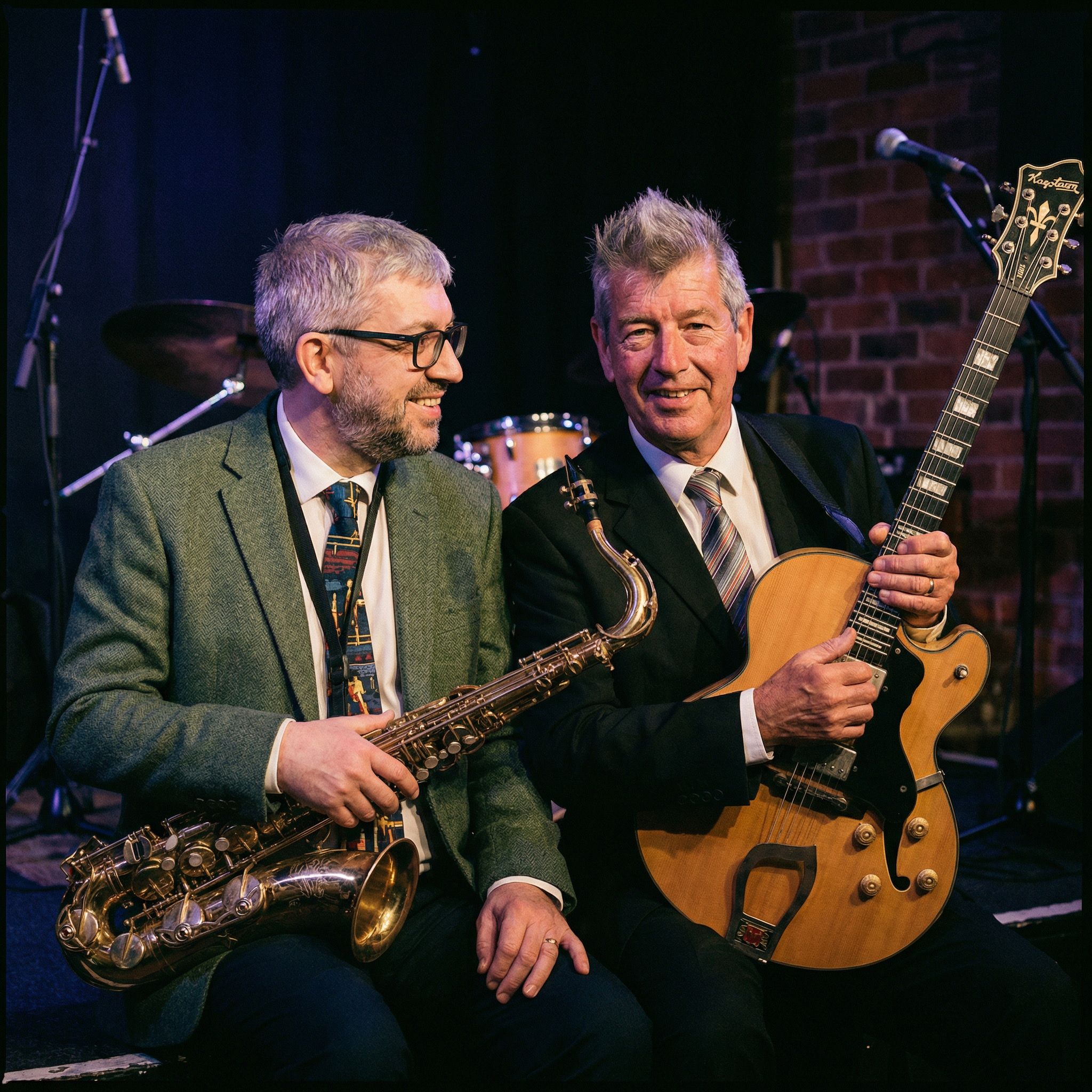 Two musicians sitting together, one holding a saxophone and the other a guitar, on stage with drums and microphones in the background.