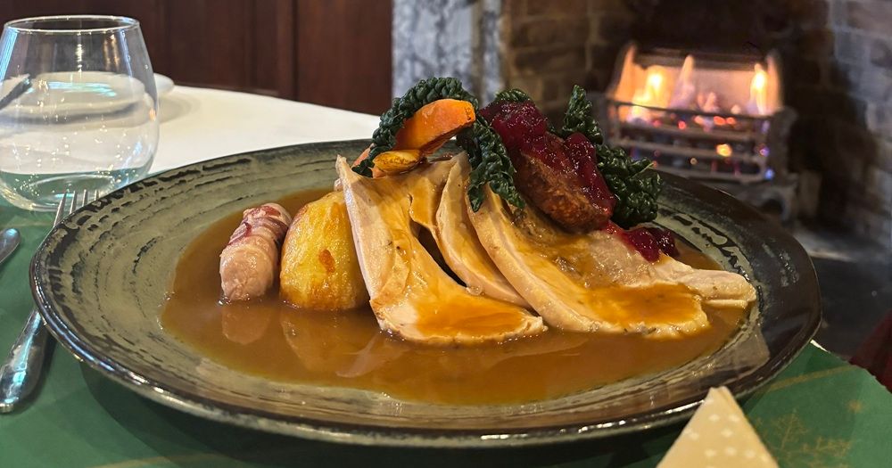 A plate of roast turkey with gravy, vegetables, roast potatoes, cranberry sauce, and pigs in blankets, set on a table near a cozy fireplace.