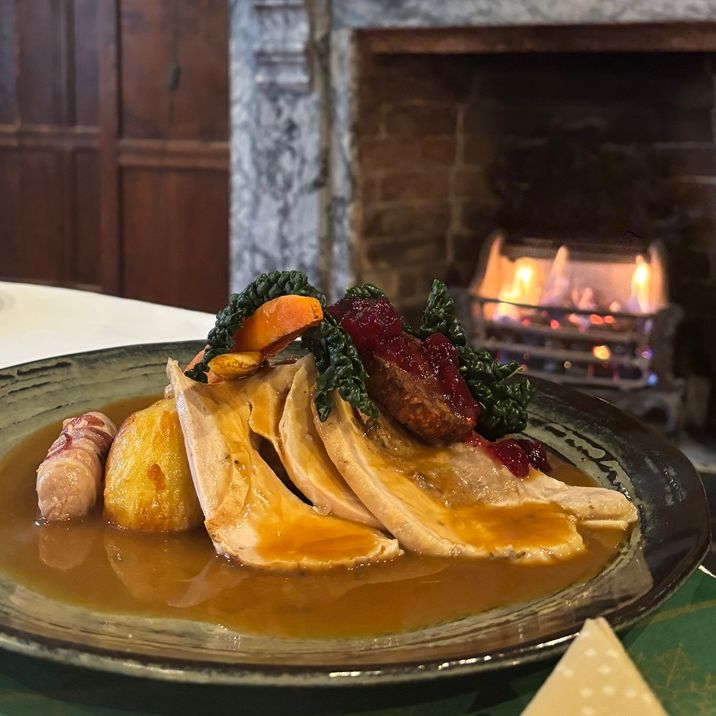 A plate of roast turkey with gravy, vegetables, roast potatoes, cranberry sauce, and pigs in blankets, set on a table near a cozy fireplace.