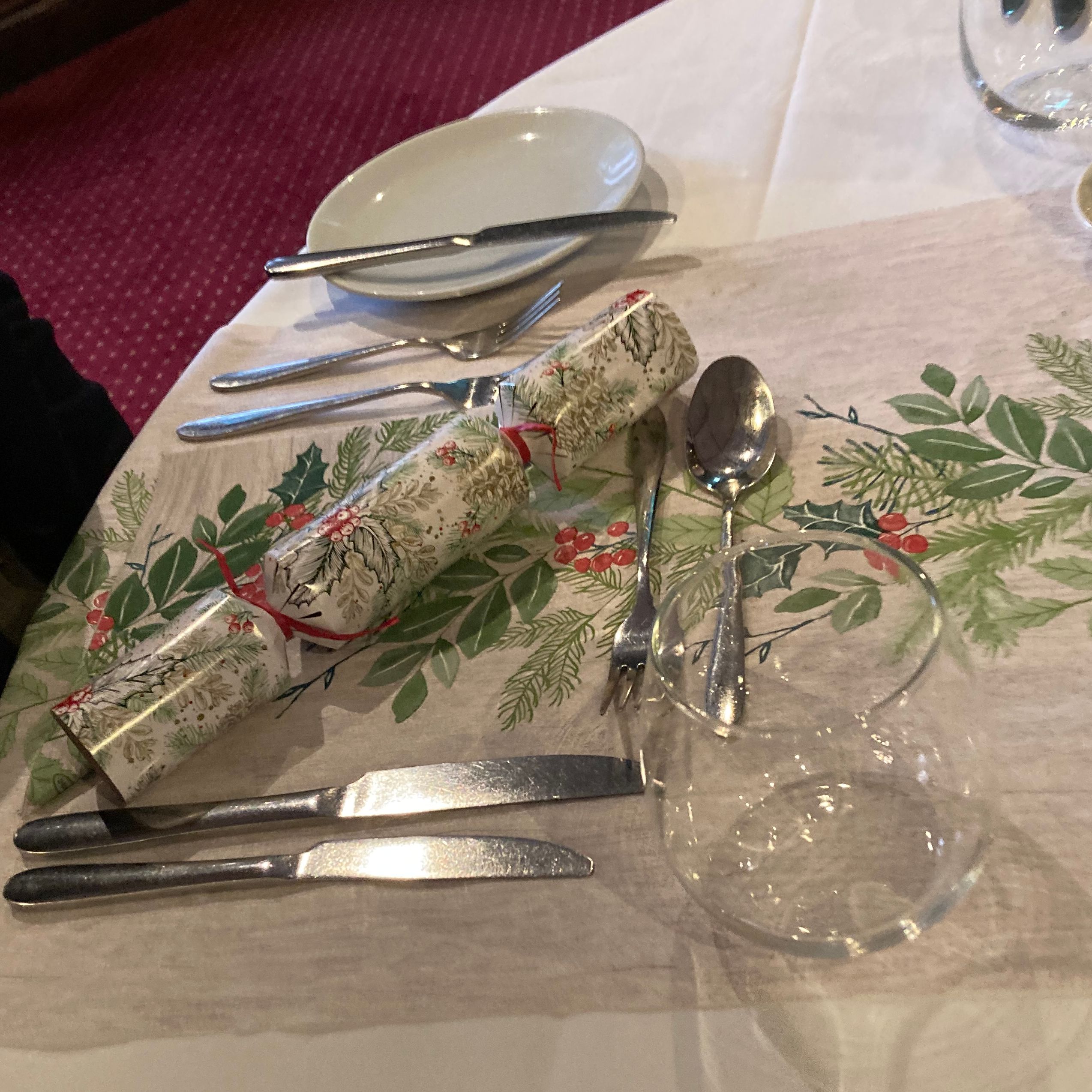 A festive table setting with a Christmas cracker, cutlery, glass, and plate on a tablecloth decorated with a holiday-themed runner.