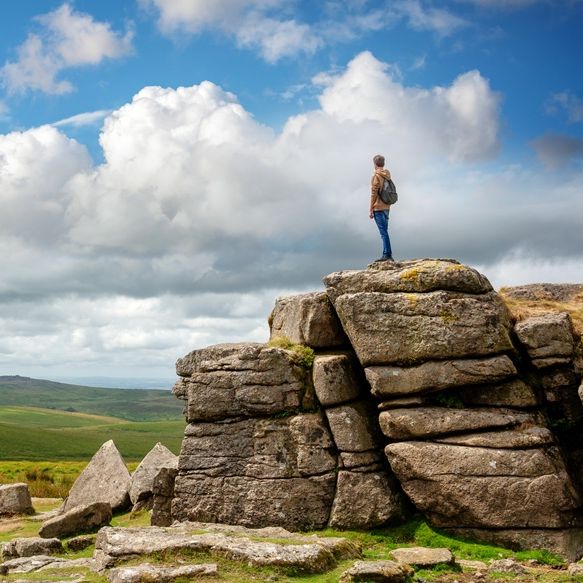 Person standing on top of a rocky tor in Dartmoor National Park, with expansive green hills and dramatic clouds in the background.