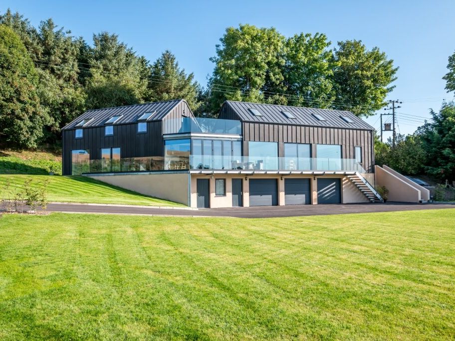 Modern house with large windows and black cladding, set on a spacious green lawn with surrounding trees