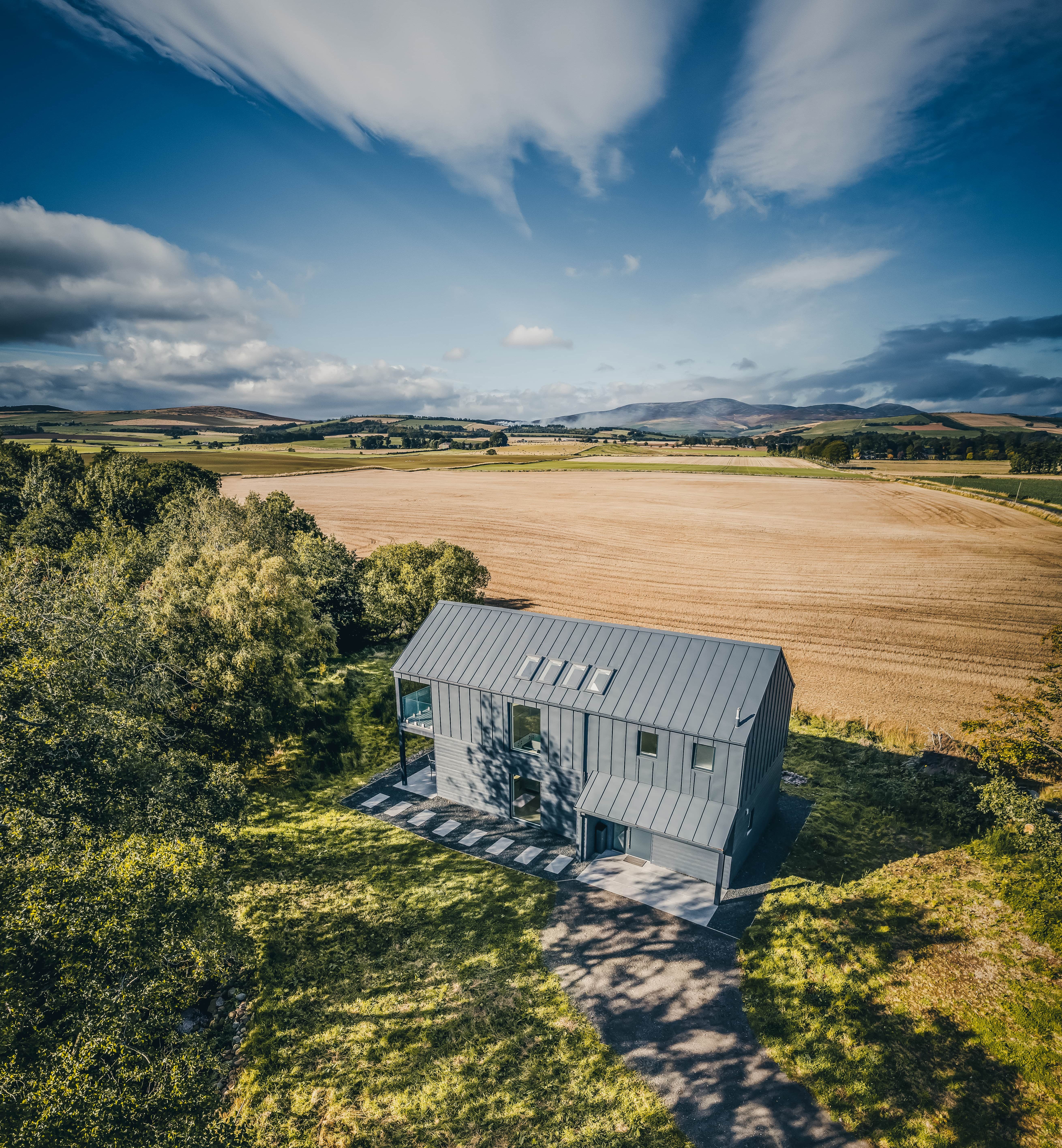 Modern house surrounded by a vast field and trees, under a dramatic sky with streaks of clouds