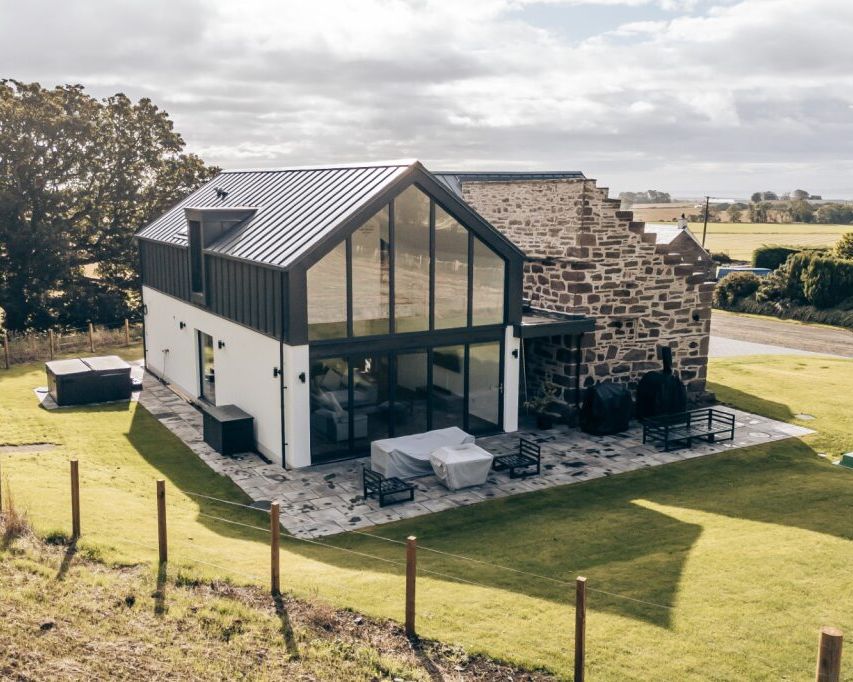 Modern farmhouse with large glass windows and stone wall exterior surrounded by green fields