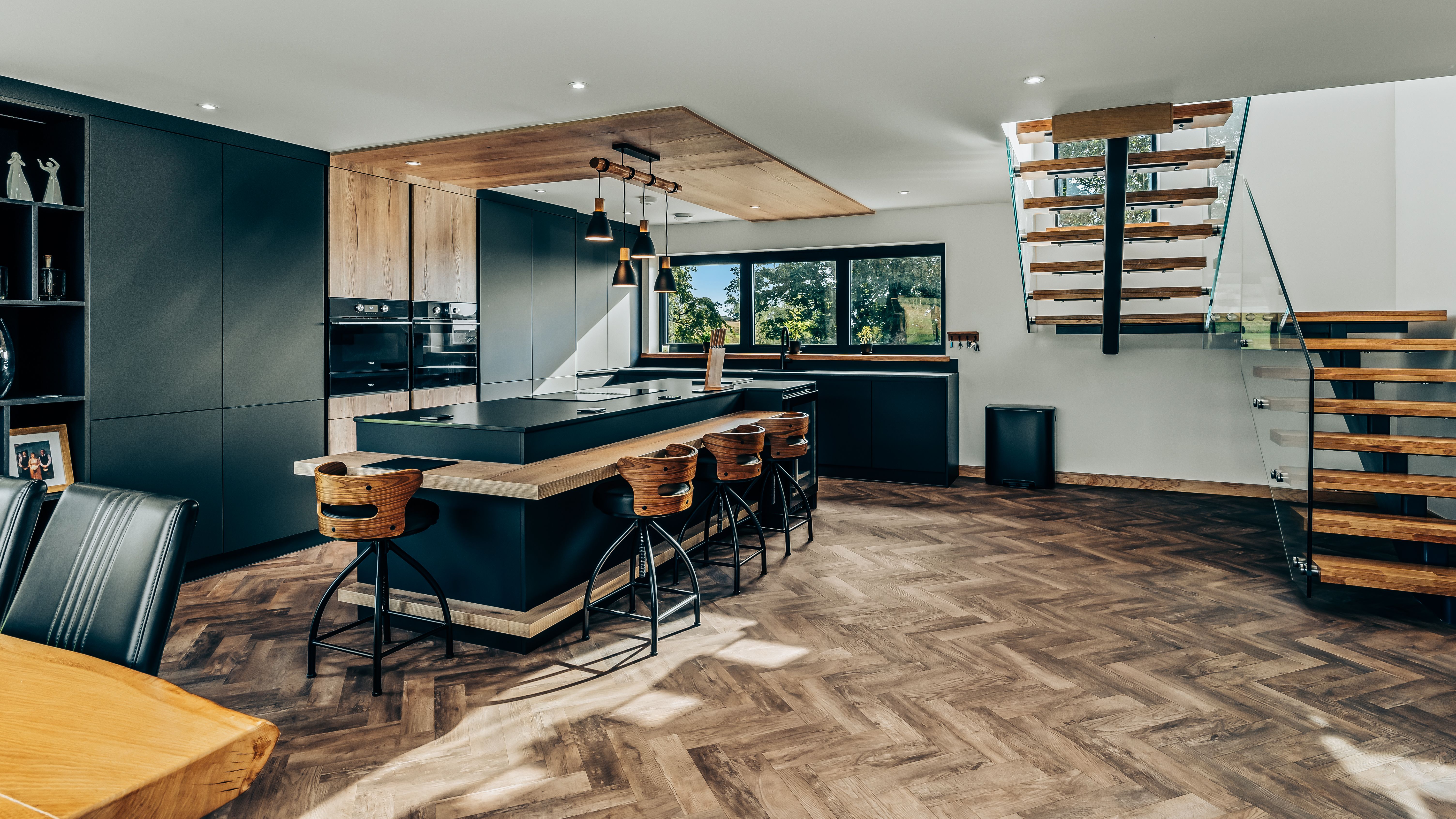 Modern kitchen with dark cabinets, wood accents, a large island with bar stools, and a floating staircase.