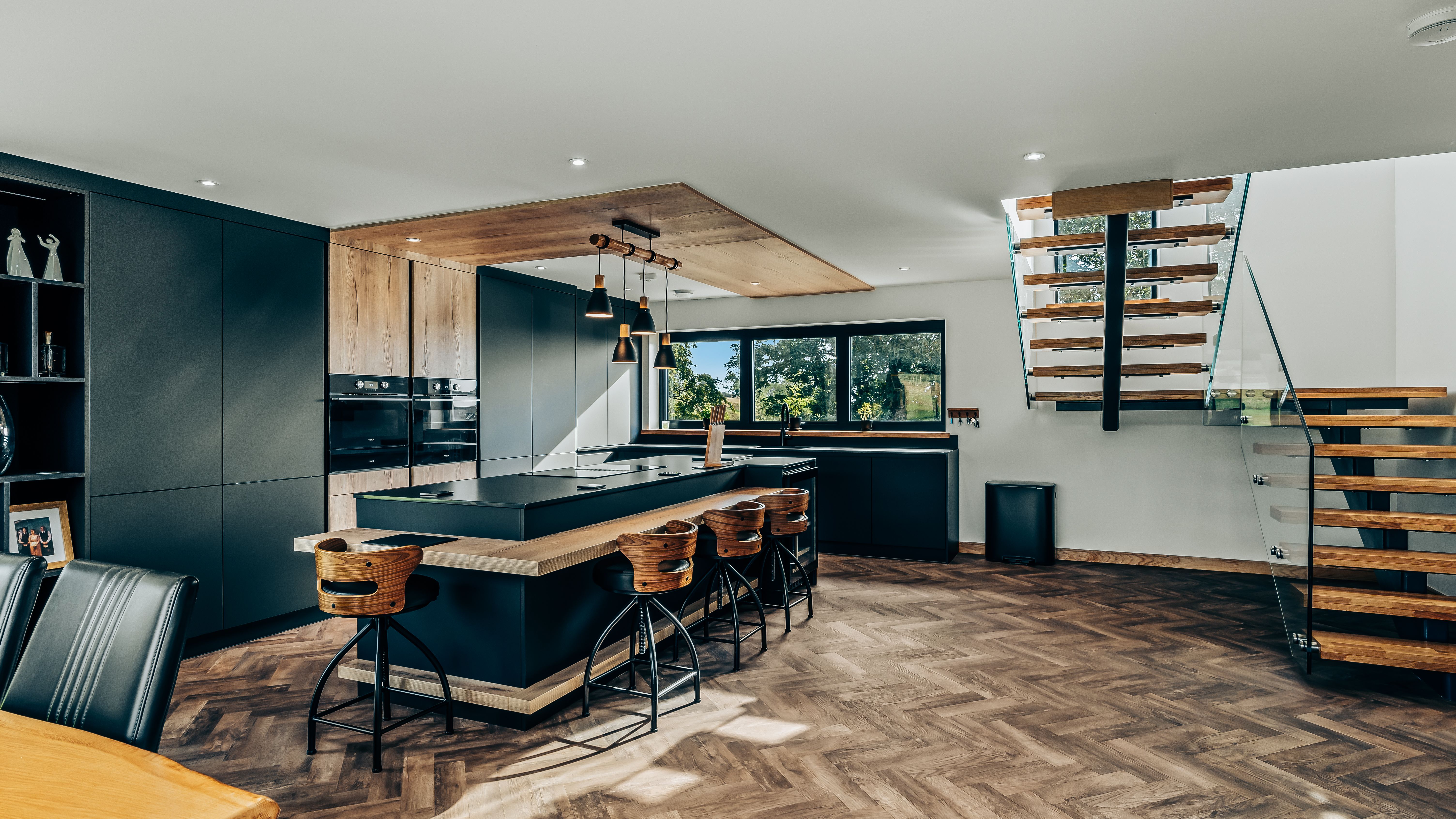Modern kitchen with dark cabinets, wood accents, a large island with bar stools, and a floating staircase.