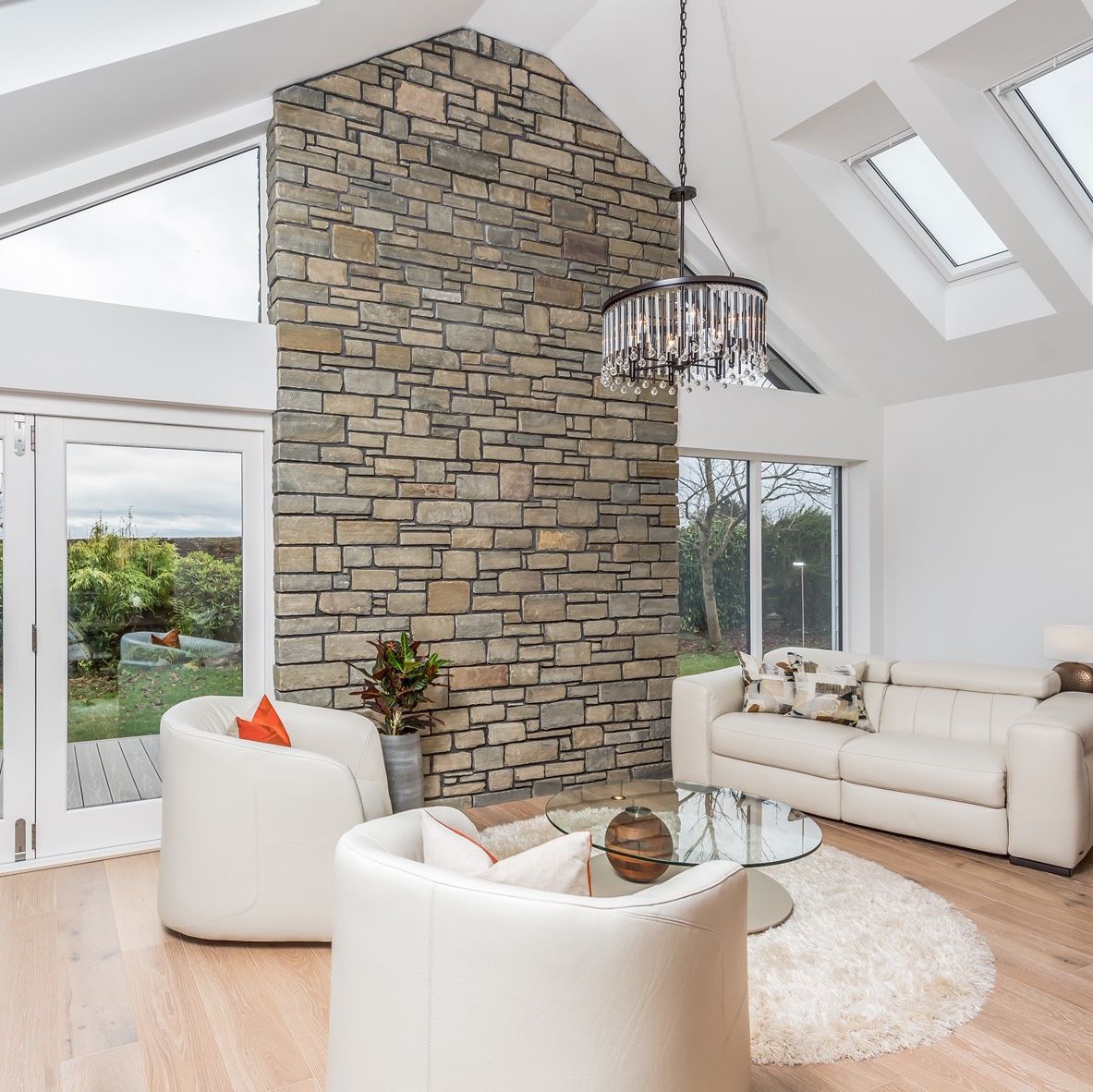 Modern living room with stone fireplace, white leather furniture, glass coffee table, and floor-to-ceiling windows.