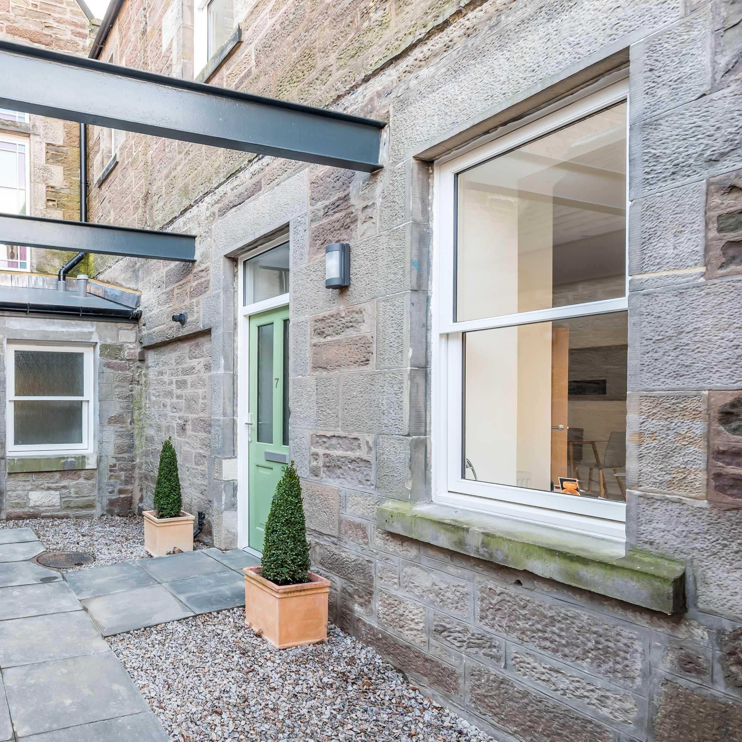 A small stone courtyard with potted topiary plants, light green doors, and large rectangular windows.